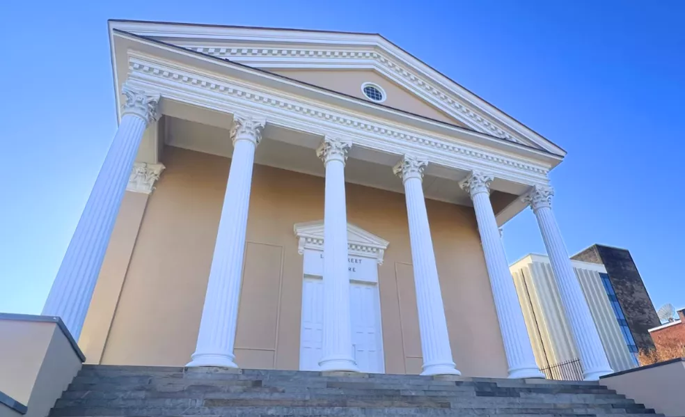 Tan stucco building with large white columns and decorative pediment. 