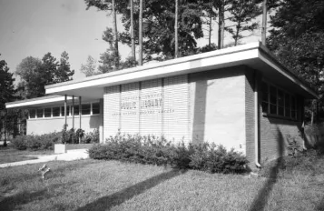 Black and white image of a midcentury building with a cantilevered roof and light colored brick, reading "Public Library" on the side of the building.  