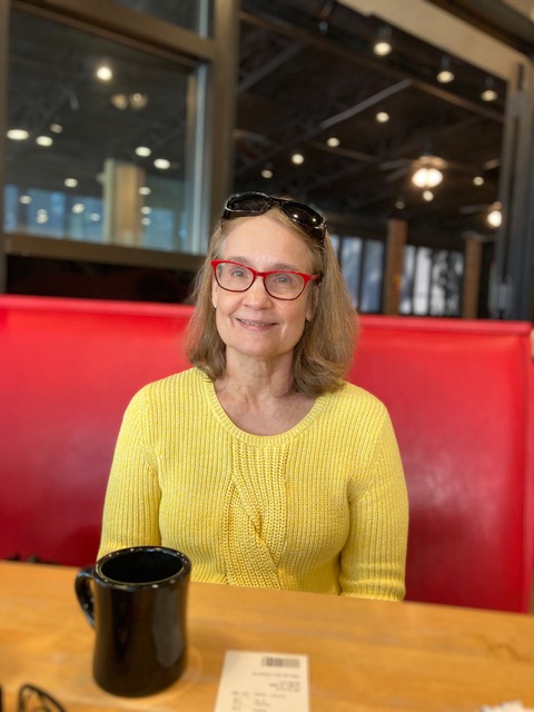 Woman in yellow shirt and red rimmed glasses sitting in a red booth.