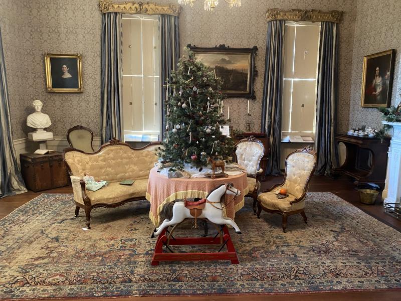 1800s house with Christmas tree on a table, surrounded by upholstered cream-colored chairs and a small couch. Rocking horse is in front of the table. 