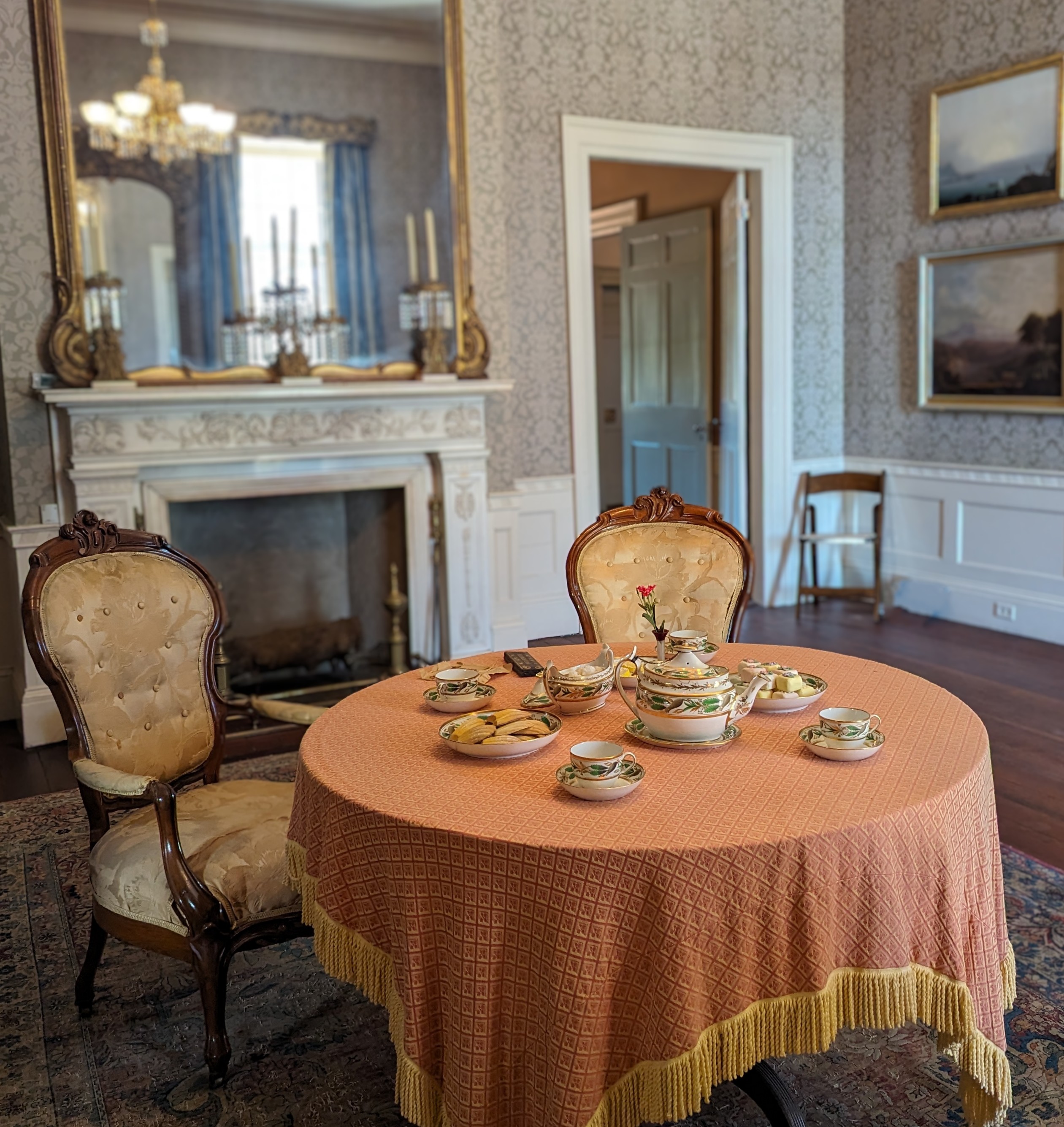 Table with fringed cloth and two chairs. On the table is an ornate tea set.