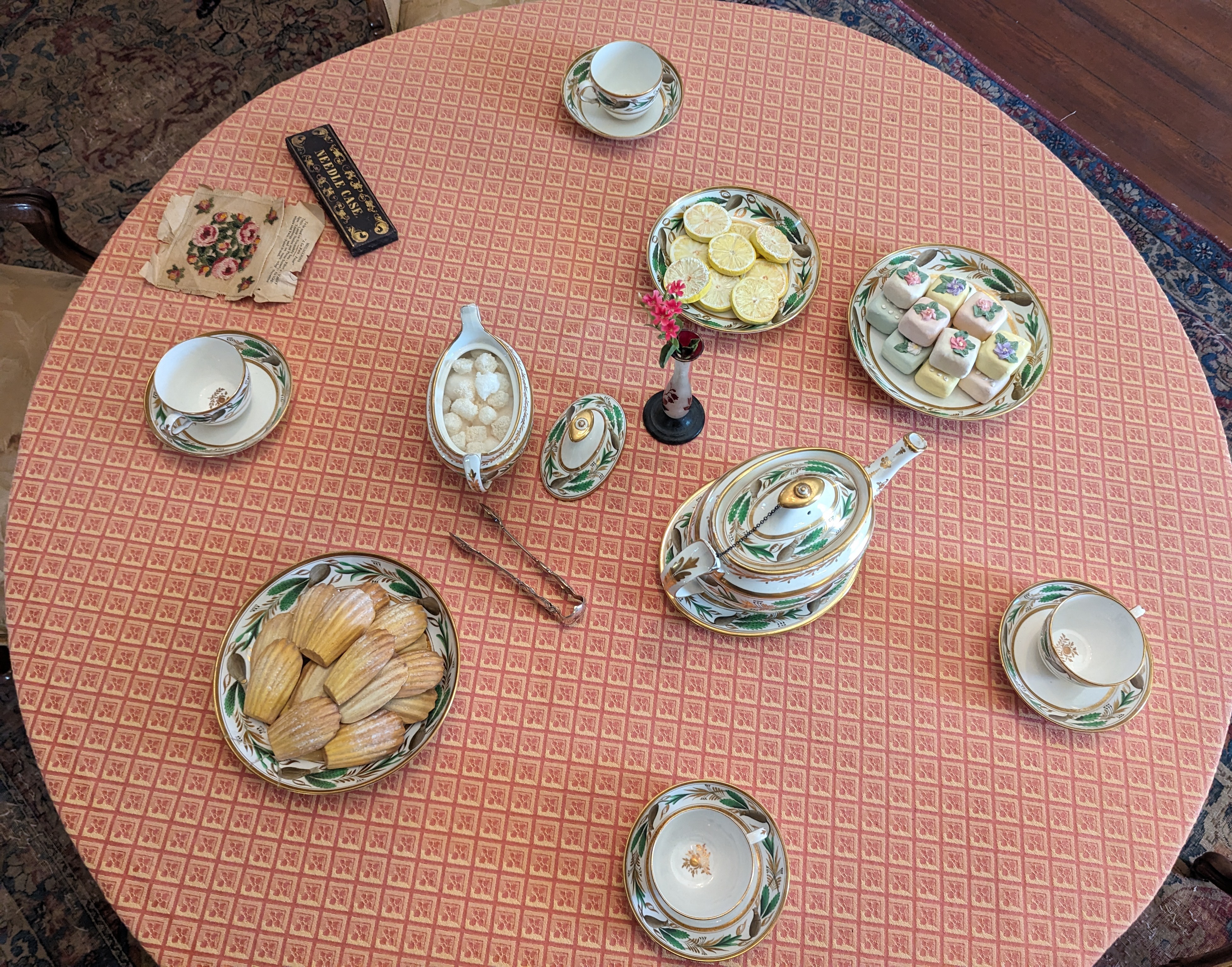 Overhead view of a tea setup with petit fours and madelines, tea cups on saucers, a teapot, sugar bowl, and creamer.